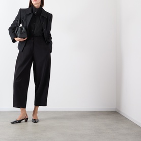 Model wearing black tailored cropped trousers with wide leg, styled with matching blazer, silk blouse and low-heel pointed shoes, carrying small
