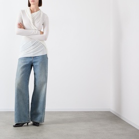 Model wearing Source Unknown Folgore Draped Top in ivory with asymmetric neckline and ruched detail, styled with wide-leg blue jeans and black heels.
