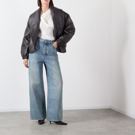 Model wearing Source Unknown Folgore Draped Top in ivory with asymmetric neckline, styled with dark bomber jacket and wide-leg blue jeans.