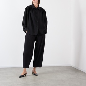 Model wearing Source Unknown Bimba Pintuck Trousers in black, wide-leg cropped style, paired with a black oversized shirt and black mules.