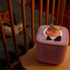 Tonies Toniebox audio player in pink, glowing ring light active, styled on wooden shelf beside a sleeping child in a cot