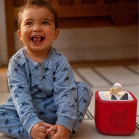 Tonies red Toniebox speaker with a Tonie figure on top, next to a smiling toddler in blue patterned pyjamas seated on a rug