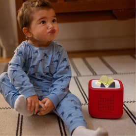 Tonies red Toniebox speaker on floor beside toddler in blue pyjamas, with a Tonie character figure on top