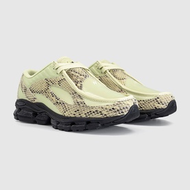 New Balance trainers in Morel Black with snake-embossed panels, pale green suede overlays, and black chunky sole. Pair shot, angled view.