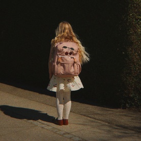 Child wearing Konges Sl&oslash;jd cherry pink backpack with character print, styled with white skirt, tights and red shoes, walking outdoors