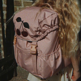 Konges Sl&oslash;jd cherry-print backpack in dusky pink with tan buckle clip, worn on back by child with long blonde hair