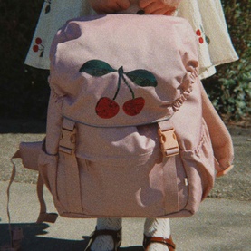 Konges Sl&oslash;jd cherry-print pink backpack with gold buckle fastenings, worn by child in white dress with red cherry print trim.