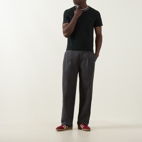 Model wearing Lacoste Crew Neck T in Dark Varech with white tipped collar, styled with charcoal trousers and red trainers.