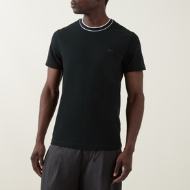 Model wearing Lacoste Crew Neck T in dark green, with contrast stripe collar and small logo chest badge, styled with dark grey trousers.