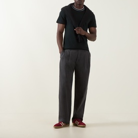 Model wearing Lacoste Crew Neck T in Dark Varech with contrast stripe collar, styled with grey trousers and red trainers.