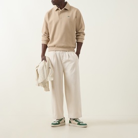 Model wearing Lacoste Viennese polo sweat with cream wide-leg trousers and green leather trainers, holding a cream jacket.