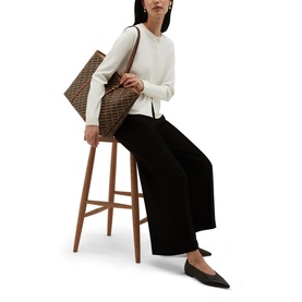Model carrying a brown monogram tote bag, styled with a cream knit top and wide-leg black trousers, seated on a wooden stool