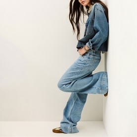 Model wearing Bash blue wide-leg jeans and denim jacket, styled with tan slides, leaning against a white wall.