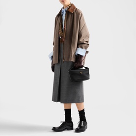 Model carrying a small black bag with gold hardware, styled with a tan jacket, grey midi skirt and black leather shoes