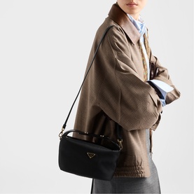 Model carrying black fabric shoulder bag with gold-tone hardware and adjustable strap, styled with a brown textured jacket and grey trousers