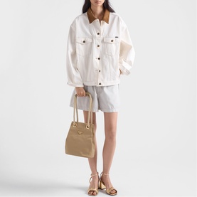 Model carrying a small beige bucket bag with gold hardware, styled with a white oversized jacket, grey shorts, and strappy sandals