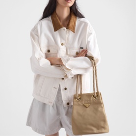 Model carrying small beige bucket bag with gold chain strap and logo hardware, styled with white jacket and grey shorts