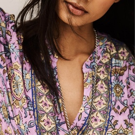 Model wearing Bash purple paisley print blouse with V-neckline, styled with a delicate gold chain necklace