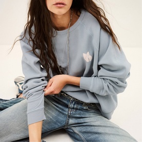 Model wearing Bash blue sweatshirt with small floral embroidery at chest, styled with light wash jeans and a delicate necklace.