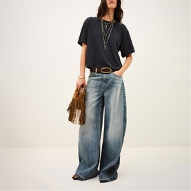 Model wearing Bash blue wide-leg jeans with dark grey oversized tee, layered necklaces and brown leather belt, carrying fringed bag.