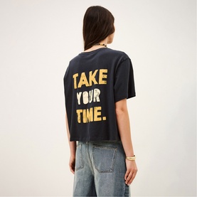 Model wearing Bash blue cropped T-shirt with gold "Take Your Time" slogan on back, styled with light wash jeans and gold bracelet.