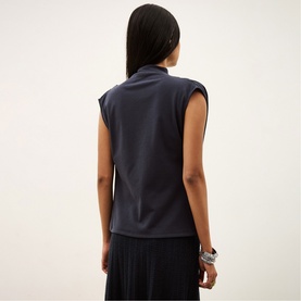 Model wearing Bash blue sleeveless top with mock neck, styled with black pleated skirt and silver cuff bracelet, back view.