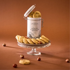 Cartwright & Butler Hazelnut & Chocolate Biscuits in a glass-lidded tin displayed on a glass cake stand with scattered hazelnuts