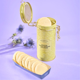 Cartwright & Butler Lemon Shortbread Biscuits in yellow hinged tin with open lid, biscuits fanned out on tray, lavender background