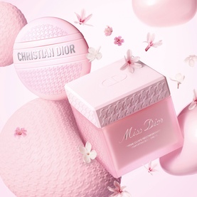 Dior Miss Dior body cream in pink embossed cube jar with matching powder puff, surrounded by cherry blossom flowers and pink spheres