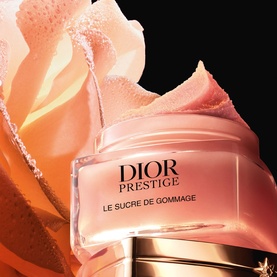 Dior Prestige Le Sucre de Gommage body scrub in open glass jar, showing pink sugar-textured formula with rose petal backdrop.
