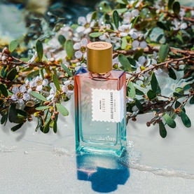 Goldfield & Banks Pacific Rock Flower EDP in teal-to-blush gradient bottle with gold cap, styled on sand with flowering foliage