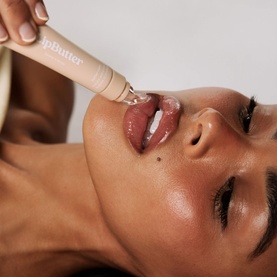 Model with Kylie Cosmetics lip butter gloss applicator being applied to lips, close-up beauty shot showing glossy nude lip finish.