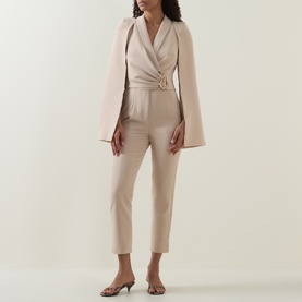 Model wearing Lavish Alice Sand jumpsuit with cape sleeves and gold buckle waist detail, styled with strappy heeled mules