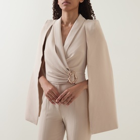 Model wearing Lavish Alice sand jumpsuit with cape sleeves and gold buckle belt detail, styled in a V-neckline silhouette.