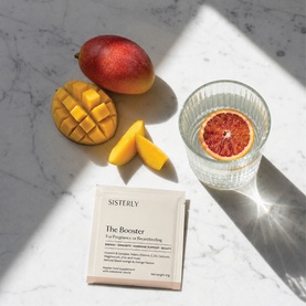 Sisterly The Booster supplement sachet on marble surface, styled with mango slices, blood orange in water glass, and sunlight shadows.