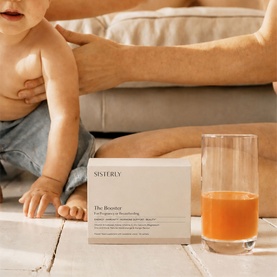 Sisterly The Booster supplement box beside a glass of orange juice, with a baby in the background on a light wooden floor.