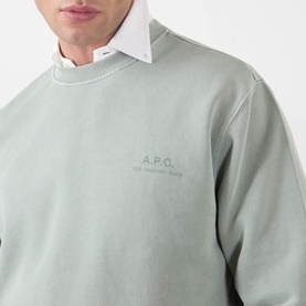 Model wearing A.P.C. Fernando Logo Sweatshirt in gray green, crew neck with tonal chest logo print, styled with white shirt collar visible.
