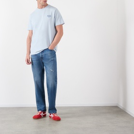 Model wearing A.P.C. light blue cotton T-shirt with small chest logo, styled with mid-wash distressed jeans and red trainers.