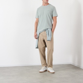 Model wearing A.P.C. gray green cotton T-shirt, styled with beige trousers and a tied shirt at the waist, white trainers.