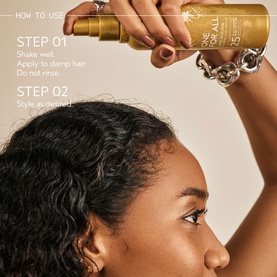 Model with Aveda treatment spray held above curly hair, step-by-step how-to-use guide overlay, gold bottle, silver chain bracelet visible