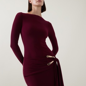 Model wearing Club London burgundy long-sleeve dress with asymmetric neckline and gold-tone buckle tie detail at hip.