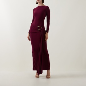 Model wearing Club London burgundy long-sleeve maxi dress with ruched waist detail and gold buckle trim, styled with gold heeled sandals.