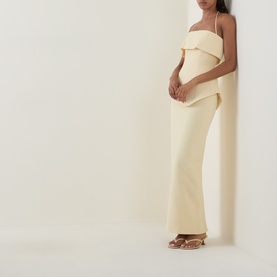 Model wearing a strapless asymmetric drape maxi dress in pale yellow with wide-leg trousers, styled with gold strappy heeled sandals.