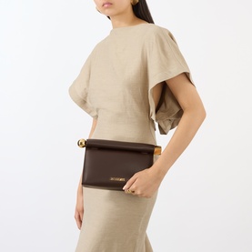 Model carrying Jacquemus midnight brown leather clutch with gold bar detail, styled with beige draped top and matching skirt