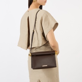 Model carrying Jacquemus Midnight Brown leather clutch with gold hardware and adjustable crossbody strap, styled with beige linen co-ord