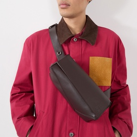 Model carrying Loewe Puzzle Featherlight Sling Bag in dark chestnut grained leather, silver-tone hardware, worn crossbody over red jacket.