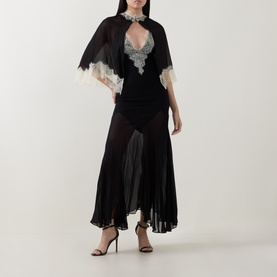 Model wearing Munthe Cielo Cape Dress in black with detachable sheer cape, lace trim, V-neckline, and midi length, styled with black heels.