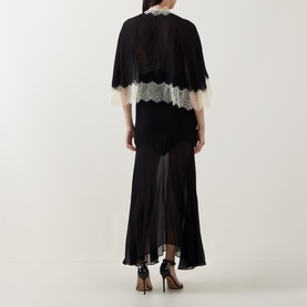 Model wearing Munthe Cielo Cape Dress in black with lace-trim detachable cape, midi-length slip dress, styled with black heels, back view.
