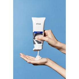Anua cleanser tube being squeezed, dispensing white foam product onto an open palm against a blue background
