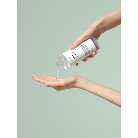 Anua toner being dispensed from a clear pump bottle onto an open palm, shown against a sage green background